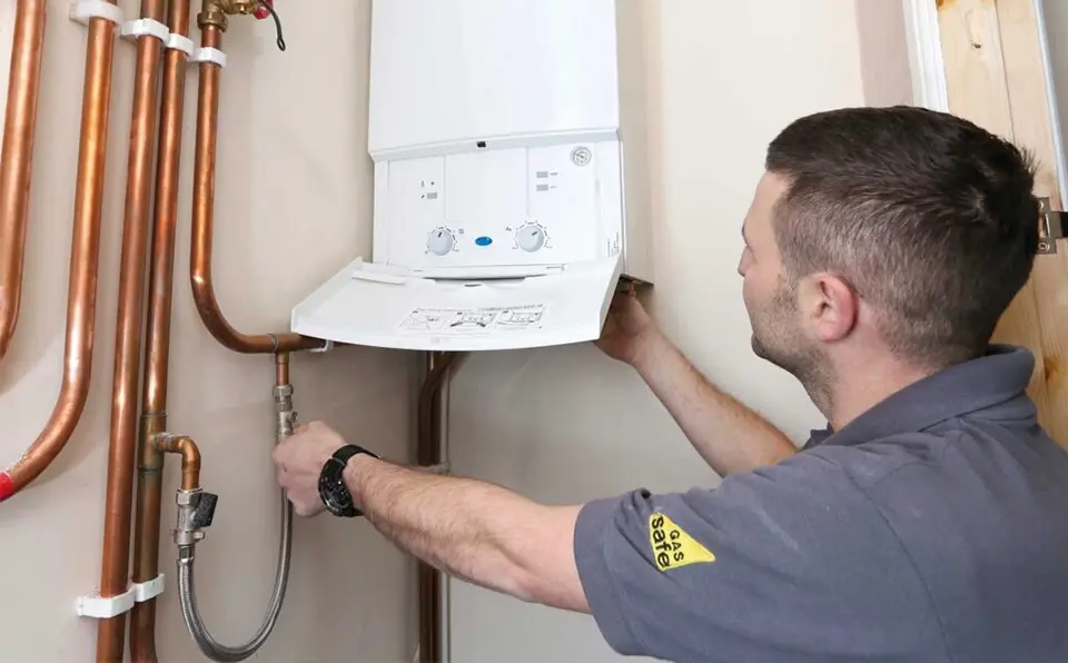 A technician adjusting a boiler unit mounted on a wall with copper pipes connected.