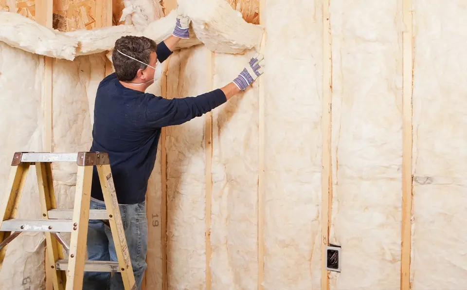 A man wearing gloves and a mask installs insulation material in a wall while standing on a ladder.