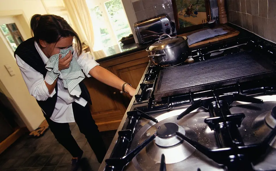 Individual covering their face while reaching for a gas stove in a kitchen setting.